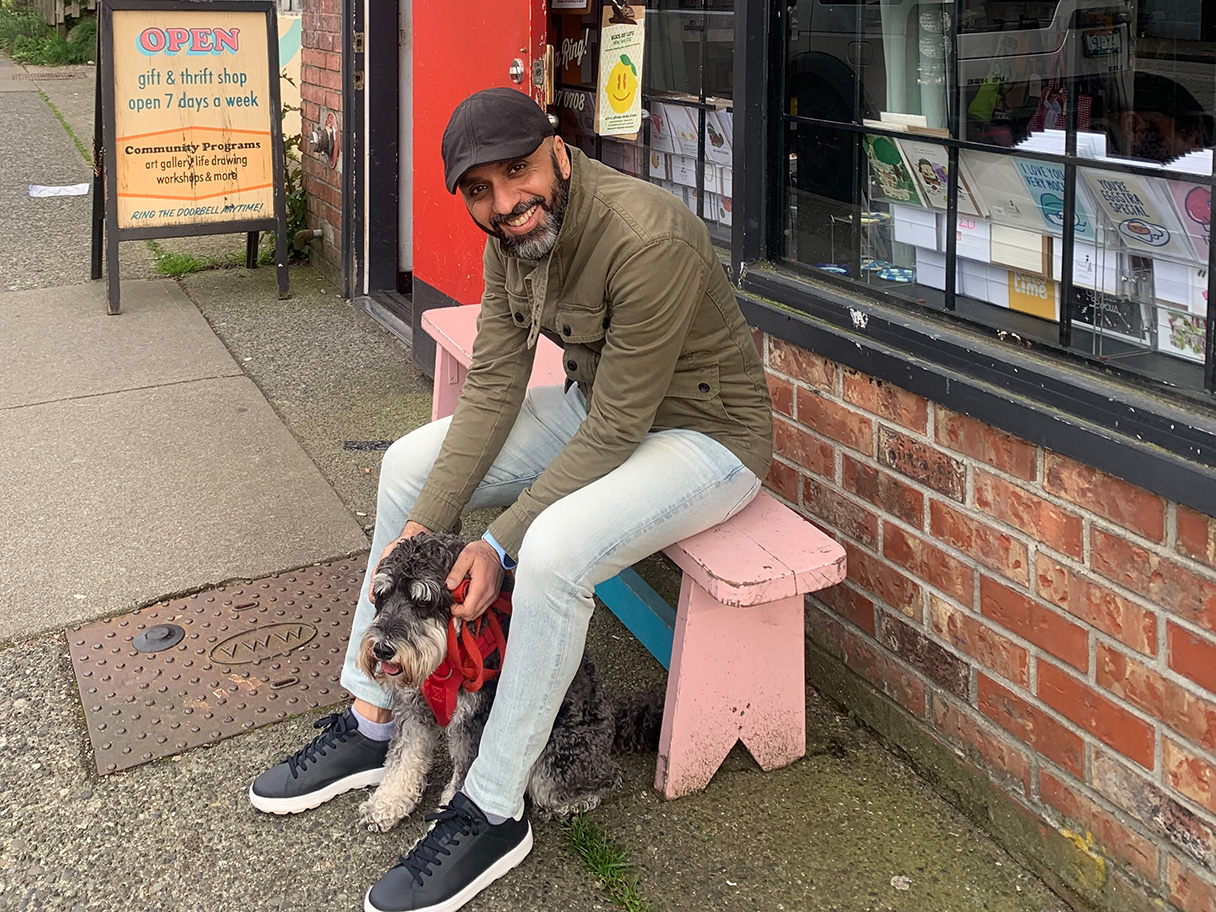 Photo of artist Ally Noormohamed sitting on the bench smiling with his dog in front of Slice of Life Gift Storefront